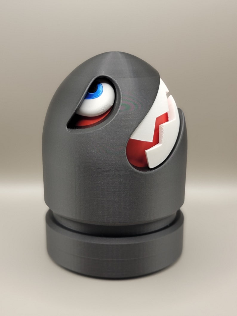 May include: A gray, 3D-printed Bullet Bill figure, a character from the Super Mario video game series. The figure has a white and red mouth, a blue and white eye, and a cylindrical base.