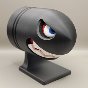 May include: A black Bullet Bill figurine with a menacing expression. The cartoon character has a white-toothed grin, red mouth, and a single eye with blue, white, and red details. The figurine is mounted on a black base.