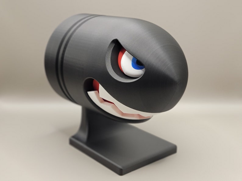 May include: A black Bullet Bill figurine with a menacing expression. The cartoon character has a white-toothed grin, red mouth, and a single eye with blue, white, and red details. The figurine is mounted on a black base.