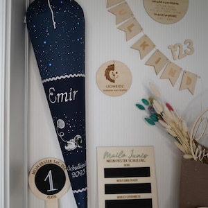 Könnte beinhalten: Eine marineblaue Schultüte mit Sternenmuster, dem Namen "Emir" und einer Astronautenillustration. Holzdekorationen, darunter ein Banner mit der Aufschrift "SCHULKIND", ein Schild mit der Aufschrift "Mein Erster Schultag" und ein Schild mit der Aufschrift "1. Klasse".