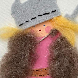 Könnte beinhalten: Eine Filzpuppe mit blonden Haaren, die einen grauen Filzhut mit einem schwarzen, gestickten Rand und ein rosa Kleid mit einem braunen Gürtel trägt. Die Puppe hat ein bemaltes Gesicht mit blauen Augen und rosa Wangen.