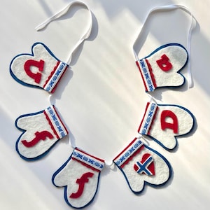 Peut inclure: Une guirlande en feutre avec des formes de mitaines blanches ornées de lettres et de motifs rouges. Chaque moufle est bordée de bleu et possède une bande décorative avec des motifs rouges, blancs et bleus. La guirlande est enfilée sur un ruban blanc.