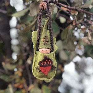 May include: A felt woodland creature ornament with a green hat and a brown acorn. The ornament is hanging from a branch.