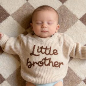 Op de afbeelding: Een beige gebreide trui met lange mouwen en de tekst Little brother geborduurd in donkerbruin garen op de borst. De trui heeft een geribbelde ronde hals en geribbelde boorden, gedragen door een slapende baby met een lichte huid en kort haar.