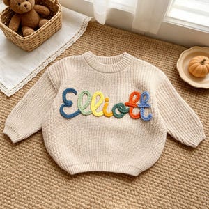 May include: A beige long-sleeve knit jumper featuring the name Elliott embroidered across the chest in raised, colourful yarn letters. Each letter is a different colour, including blue, green, yellow, orange, and purple, creating a textured design.