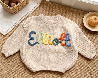 Könnte beinhalten: Ein beigefarbener, langärmeliger Strickpullover mit dem Namen Elliott, der in erhabenen, bunten Garnbuchstaben auf die Brust gestickt ist. Jeder Buchstabe hat eine andere Farbe, darunter Blau, Grün, Gelb, Orange und Violett, was ein strukturiertes Design ergibt.