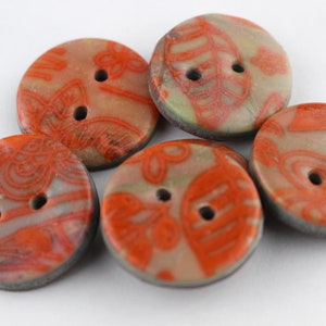 Può includere: Cinque bottoni rotondi a due fori con un motivo floreale grigio e arancione. I bottoni sono in ceramica e hanno un bordo leggermente rialzato.