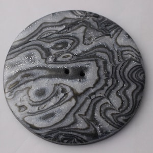 Puede incluir: Un botón redondo gris y blanco con un patrón de mármol en espiral. El botón tiene dos agujeros en el centro.
