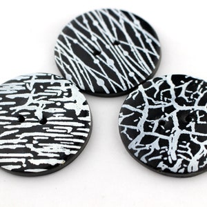 Op de afbeelding: Drie zwart-witte decoratieve knopen met een ronde vorm. Elke knoop heeft een uniek wit patroon, waaronder lijnen en een webachtig ontwerp. De knopen hebben twee gaten om op te naaien.