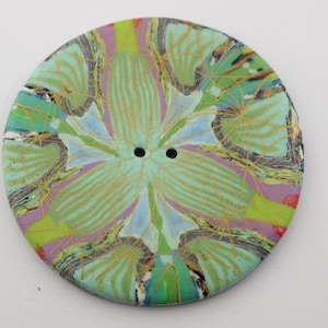 May include: A large, round button with a colorful floral design. The button features a pattern of green, blue, purple, and yellow hues, with two small holes in the center. The button is approximately 2 inches in diameter.