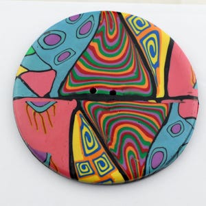 Puede incluir: Un botón redondo y colorido con un diseño geométrico. El botón está hecho de arcilla polimérica y presenta una variedad de colores, incluyendo rosa, azul, verde, amarillo y naranja. El botón tiene dos agujeros en el centro.