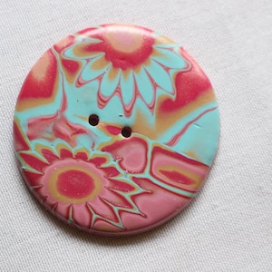 Puede incluir: Un botón grande y redondo con un diseño floral. El botón presenta una mezcla de colores, incluyendo rosa, azul claro y amarillo. Tiene dos agujeros para coser. El botón es probablemente de arcilla polimérica.