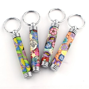 Op de afbeelding: Vier kleurrijke sleutelhangers met zilveren sleutelringen. Elke sleutelhanger heeft een cilindrische hanger met een ander bloemen- en dierenontwerp. De hangers zijn gemaakt van polymer clay en hebben een glanzende afwerking.