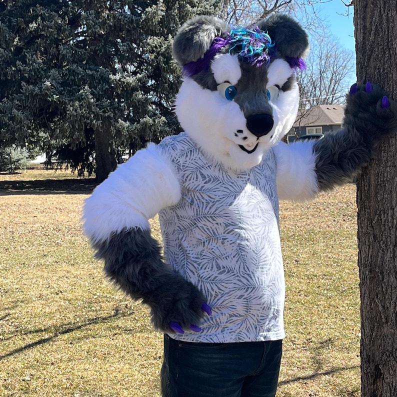 May include: A gray and white furry wolf costume with a purple and blue mohawk. The wolf is wearing a white shirt with a gray and white leaf pattern. The wolf is standing next to a tree.