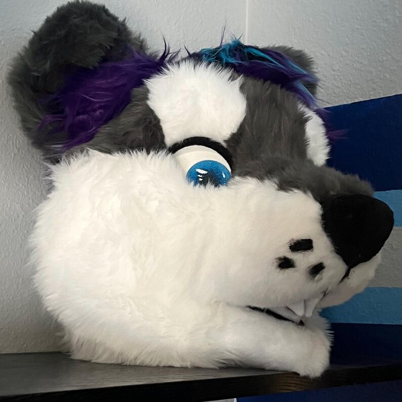 May include: A furry gray and white animal head with blue eyes and purple and blue fur accents. The head is tilted to the side and has a toothy grin.