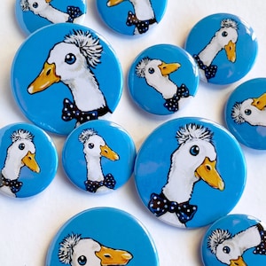 May include: A set of ten blue and white round buttons featuring cartoon illustrations of ducks with a mohawk hairstyle wearing bow ties. The ducks are on a bright blue background.