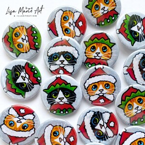 May include: A collection of 13 round pinback buttons featuring hand-painted illustrations of cats wearing festive holiday attire. The cats are depicted in various poses, some wearing Santa hats, others elf hats, and some with both. The buttons are a mix of black and white cats and orange tabby cats. The background is a light gray.