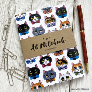 May include: A white A6 notebook with a brown paper cover that says "A6 Notebook - 48 Lined Pages". The cover is decorated with a pattern of cartoon cats wearing bow ties. The cats are in various colours, including black, white, grey, orange, and brown.