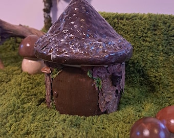 May include: A ceramic mushroom house with a dark brown, textured roof and a wooden door. The house is set amongst small mushroom figurines on a green, moss-like surface. A sign that says "FARYLAND" is in the background.