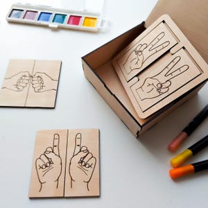 May include: A set of wooden hand gesture cards in a wooden box. The cards feature different hand gestures, including a fist bump, peace sign, and a single finger pointing up. The box also includes a set of markers for coloring the cards.