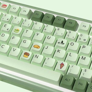 May include: A green keyboard with light green keys and various food-themed keycaps, including a burger, cookies, a cake, a teacup, and an ice cream cone. The keys are labeled with letters, numbers, and symbols.