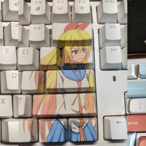 May include: A white keyboard with custom keycaps featuring anime characters. The keycaps are printed with images of two female anime characters, one with blonde hair and a blue dress and the other with black hair and a blue jacket. The keyboard has a light blue and pink colour scheme.