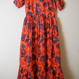 May include: An orange floral print dress with short sleeves and a square neckline. The dress features a fitted bodice and a flowing skirt with a ruffled hem. The floral pattern includes purple and teal accents.