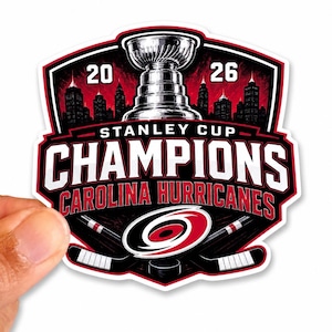 May include: A die-cut sticker featuring a black, red, and white graphic of the Stanley Cup, a city skyline, and two crossed hockey sticks. The text reads 20 26 STANLEY CUP CHAMPIONS CAROLINA HURRICANES, with the team's hurricane logo centered below.