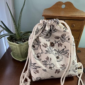 Könnte beinhalten: Ein Rucksack mit Kordelzug und floralem Muster in Grau- und Weißtönen. Die Tasche hat eine kleine Tasche mit Holzknopfverschluss. Der Kordelzug besteht aus dickem, cremefarbenem Seil.