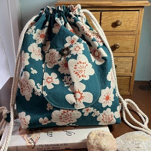 May include: A teal drawstring backpack featuring a white and orange floral pattern. The bag has a front pocket secured with a single brown button and thick white rope straps that cinch the top opening closed.