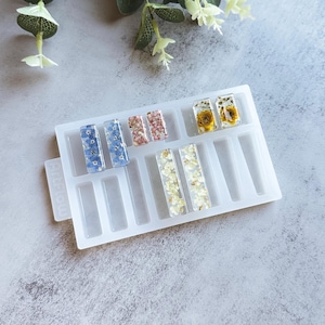 May include: A white silicone mold with 10 rectangular cavities for making resin earrings. The mold has a label that says "Make It Glow". The cavities are filled with dried flowers and leaves.