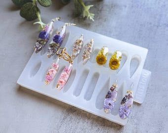 May include: A white silicone mold with 10 teardrop-shaped cavities for making resin earrings. The mold is filled with finished resin earrings with dried flowers and gold flakes. The earrings are in shades of purple, pink, and yellow.