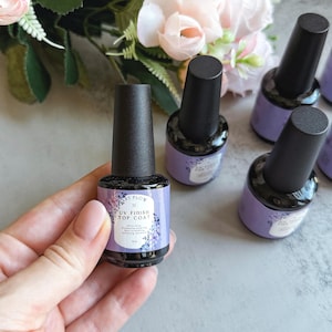Peut inclure: Cinq flacons de vernis à ongles transparent avec des bouchons noirs. Les flacons ont une étiquette violette et blanche avec le texte "UV Finish Top Coat" et "Art Flow".