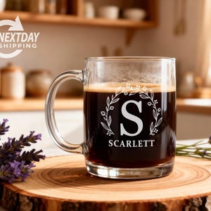 May include: A clear glass mug filled with dark liquid, possibly coffee, featuring a white floral wreath design with the letter "S" and the name "Scarlett". The mug sits on a wooden coaster, with lavender sprigs nearby.