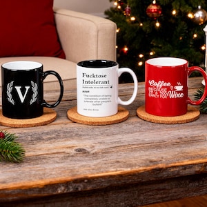 Pu&ograve; includere: Tre tazze in ceramica su sottobicchieri in sughero. Una tazza nera con una "V" bianca e un design a corona d'alloro, una tazza bianca con la scritta "Fucktose Intolerant" e una tazza rossa con la scritta "Coffee because it's too early for Wine".