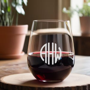 Peut inclure: Un verre &agrave; vin en verre transparent avec un monogramme des lettres "AHW" grav&eacute; sur le c&ocirc;t&eacute;. Le verre est rempli de vin rouge et repose sur un dessous de verre en bois.