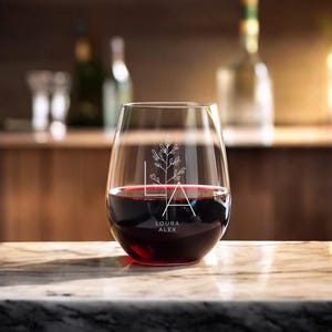 Peut inclure: Un verre &agrave; vin sans pied transparent avec un motif floral et le texte "Laura Alex" grav&eacute; sur le c&ocirc;t&eacute;. Le verre est rempli de vin rouge.