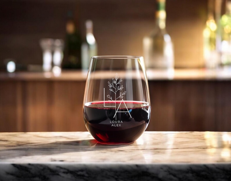 Peut inclure: Un verre &agrave; vin sans pied transparent avec un motif floral et le texte "Laura Alex" grav&eacute; sur le c&ocirc;t&eacute;. Le verre est rempli de vin rouge.