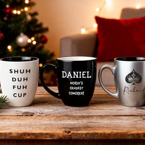 Pu&ograve; includere: Tre tazze in ceramica con interni neri su una superficie di legno. Una &egrave; bianca con le parole "SHUH DUH FUH CUP". Un'altra &egrave; nera con "DANIEL WORLD'S OKAYEST COWORKER". La terza &egrave; argentata con un disegno di picche e il nome "Richard".