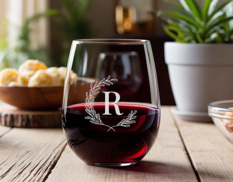 Peut inclure: Un verre &agrave; vin sans pied en verre transparent rempli de vin rouge. Le verre est d&eacute;cor&eacute; d'un motif grav&eacute; blanc repr&eacute;sentant une couronne avec la lettre "R" au centre.