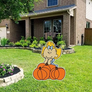May include: A yard sign featuring a cartoon character with blonde hair and a blue bow sitting atop a cluster of three orange pumpkins. The character has a smiling expression with hands resting under the chin, rendered in a flat, graphic illustration style.