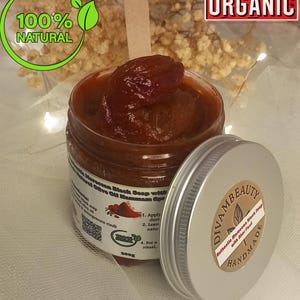 Puede incluir: Un tarro de jabón negro marroquí con un aplicador de madera. El tarro está abierto, revelando una pasta marrón rojiza oscura. La etiqueta dice "Authentic Moroccan Black Soap with Argan Oil". La imagen incluye el texto "100% Natural" y "Organic".