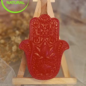 May include: A red Hamsa hand-shaped soap with intricate details, displayed on a small wooden easel. The soap is a vibrant red color and features a detailed floral design. The image includes the text "100% NATURAL" and "ORGANIC".