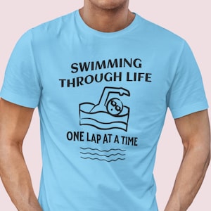 May include: A light blue t-shirt with a black graphic of a person swimming with the text "Swimming Through Life One Lap At A Time".