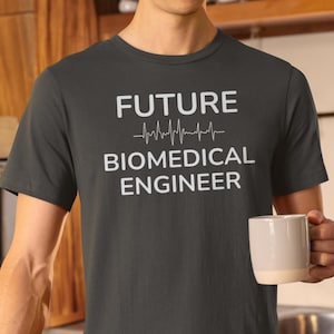 Könnte beinhalten: Dunkelgraues T-Shirt mit dem Text "FUTURE" über einer Herzschlag-Grafik, gefolgt von "BIOMEDICAL ENGINEER" in Weiß. Eine Person hält einen weißen Becher. Das Shirt besteht aus einem weichen Material.