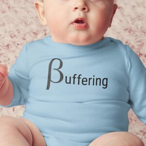 May include: A light blue baby onesie with long sleeves. The word "Buffering" is printed on the front in black, with the Greek letter beta preceding it. The baby is sitting on a pink patterned surface.