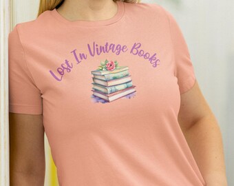 Może przedstawiać: Różowy T-shirt z napisem "Lost In Vintage Books" w fioletowym kolorze. Pod napisem znajduje się ilustracja stosu starych książek z różowym kwiatem na górze.