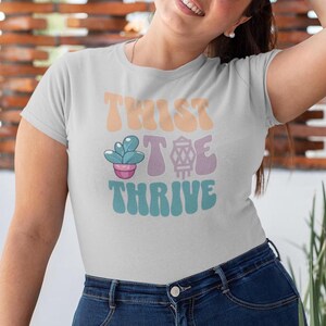 Könnte beinhalten: Ein weißes T-Shirt mit dem Text "Twist Toe Thrive" in einer Retro-Schrift. Der Text ist in den Farben Pfirsich, Lila und Türkis gehalten. Zwischen den Wörtern "Toe" und "Thrive" befindet sich eine kleine, karikaturhafte Sukkulente in einem rosafarbenen Topf.