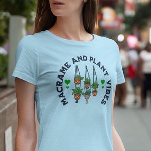Könnte beinhalten: Hellblaues T-Shirt mit einem Grafikdruck von drei hängenden Pflanzen in Makramee-Pflanzenhängern. Der Text "Macrame and Plant Vibes" ist in einem Kreis um die Pflanzen geschrieben.