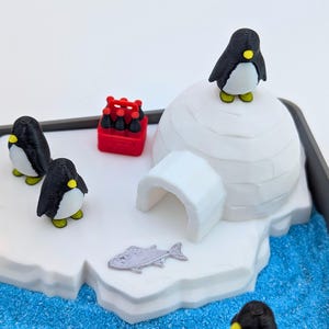 May include: A miniature arctic scene featuring a white igloo, three black and white penguins with yellow accents, and a red crate of bottles. A silver fish and blue textured base complete the diorama.